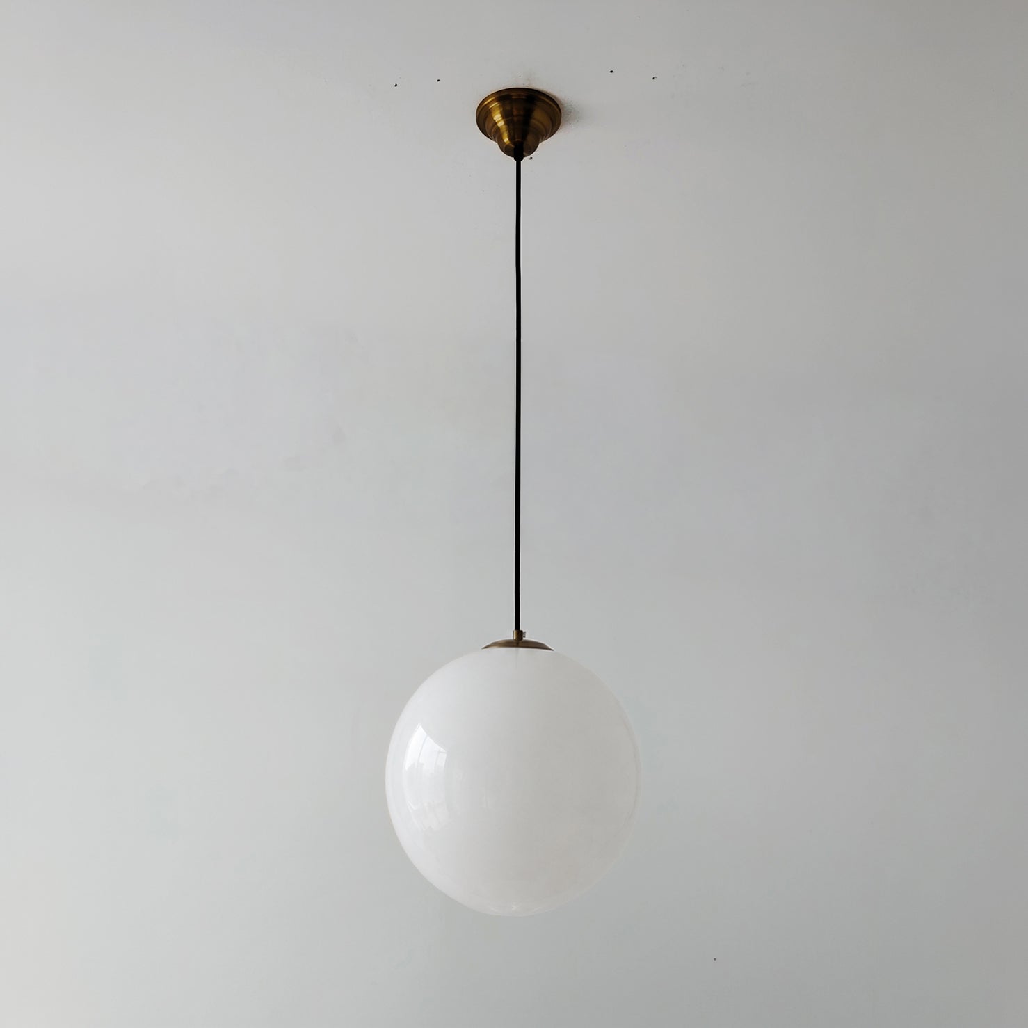 Unlit White mid century style glass globe pendant light with a brass ceiling canopy and black cord against a plain wall.