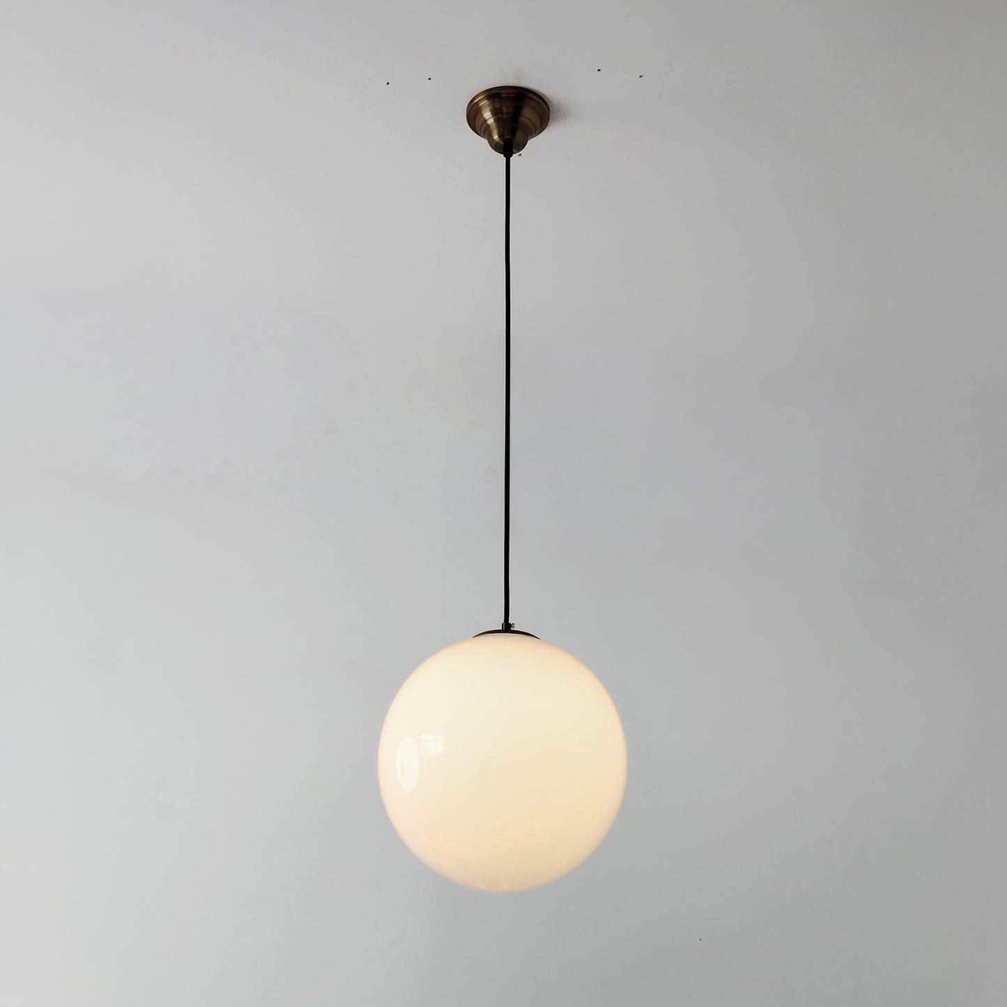Lit White mid century style glass globe pendant light with a brass ceiling canopy and black cord against a plain wall.