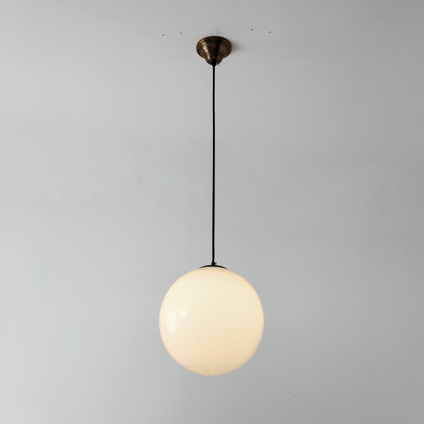 Lit White mid century style glass globe pendant light with a brass ceiling canopy and black cord against a plain wall.